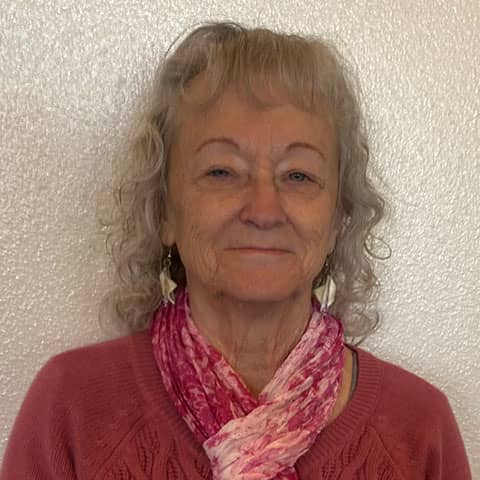 Elderly woman with curly blonde hair, wearing a red sweater and pink scarf, smiling gently at the camera.