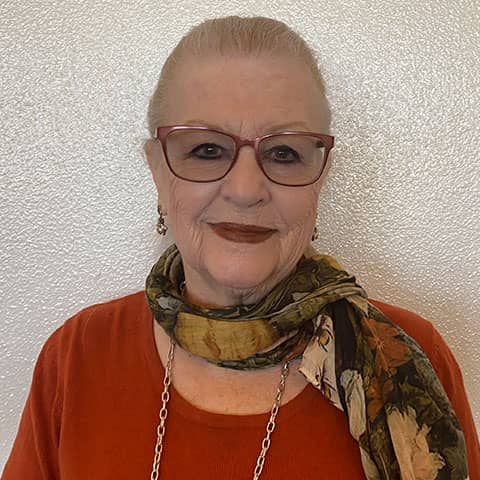 Elderly woman smiling, wearing red glasses, an orange shirt, and a green floral scarf. she has short white hair and a silver necklace.