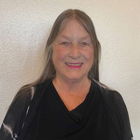 Portrait of an elderly woman with long gray hair and a black top, smiling at the camera against a white wall background.