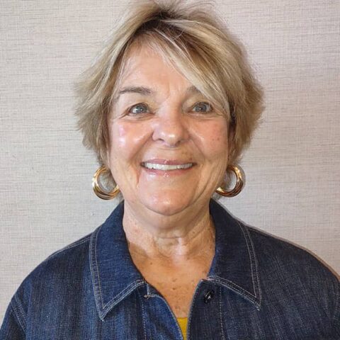 An older woman with short blonde hair and hoop earrings, smiling in front of a plain beige background, wearing a denim shirt.