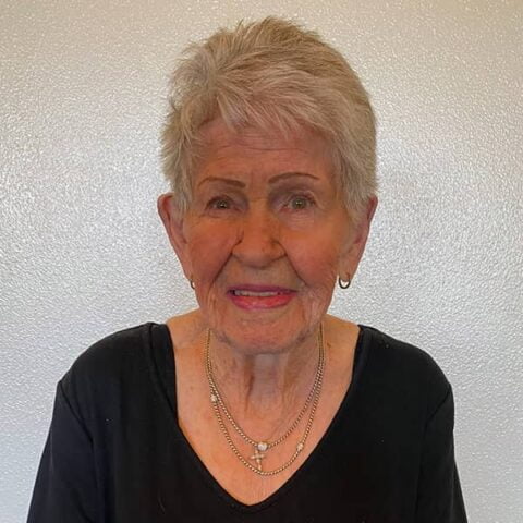 A portrait of an elderly woman with short gray hair and earrings, smiling at the camera, wearing a black top and a silver necklace.