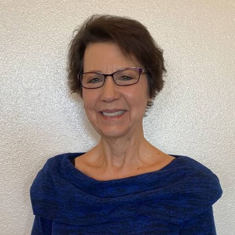 A smiling elderly woman with short brown hair, wearing glasses and a blue cowl neck sweater, stands against a plain white wall.