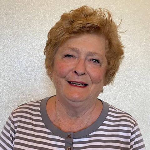 Smiling elderly woman with short reddish hair, wearing a striped top, posing against a white wall.