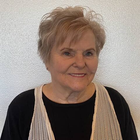 Elderly woman with short brown hair and a gentle smile, wearing a black top and white scarf, against a white background.