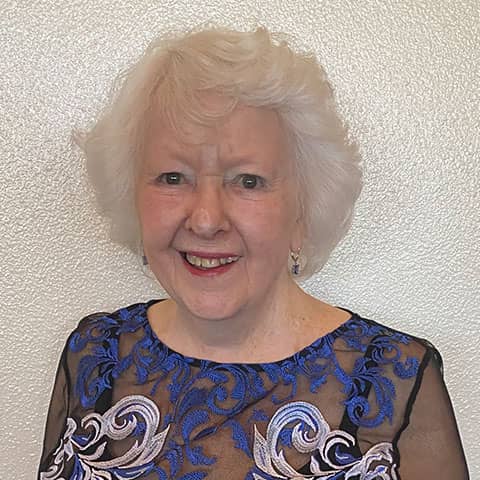 An elderly woman with white hair smiles at the camera, wearing a blue and silver floral lace dress against a white background.