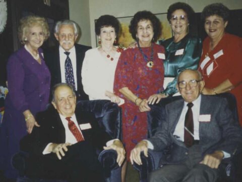 Group of seven older adults smiling at a social gathering, four women and three men, dressed in semi-formal attire with name tags.