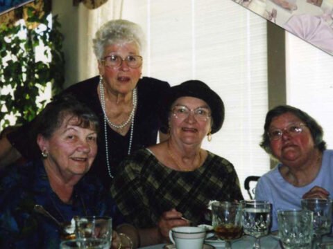 Four elderly women enjoying a meal together at a table, smiling towards the camera.