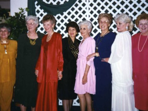Eight elderly women in formal attire posing together in front of a lattice backdrop.