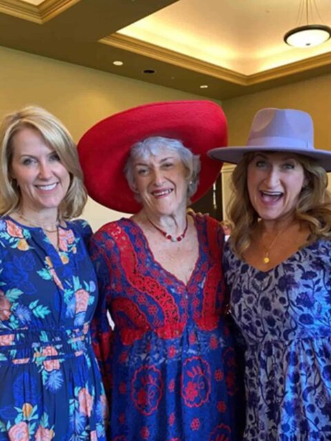 Three smiling women standing together, two in floral dresses and hats, the middle one in a red hat and blue dress.