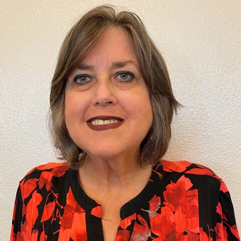 Middle-aged woman with shoulder-length brown hair and a red floral blouse smiling at the camera against a plain white background.