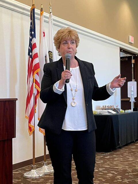 A woman with short blonde hair speaks into a microphone while standing in a conference room with flags in the background.