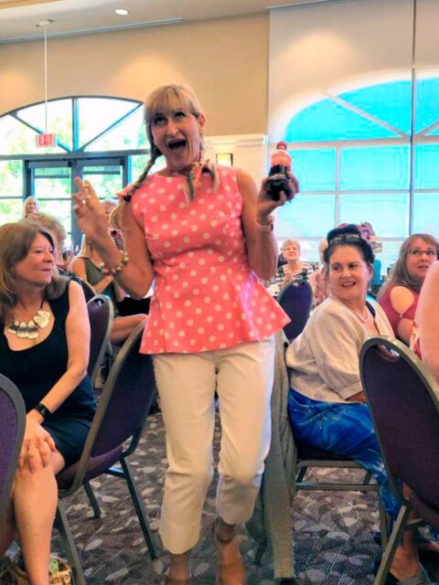 Woman in a pink polka dot blouse and white pants stands excitedly in a crowd, holding a bottle and making a surprised facial expression.