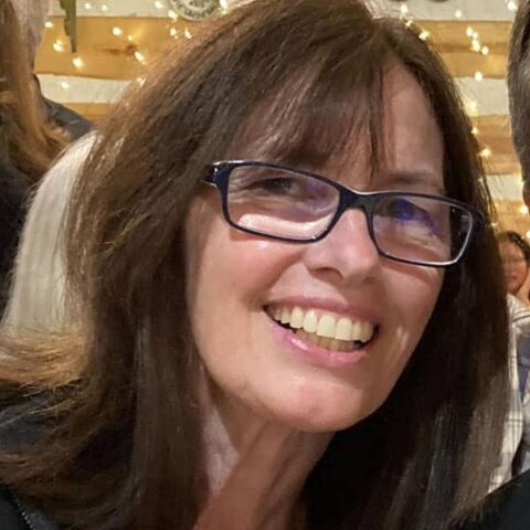 A smiling woman with long brown hair and glasses, standing in a room with string lights in the background.