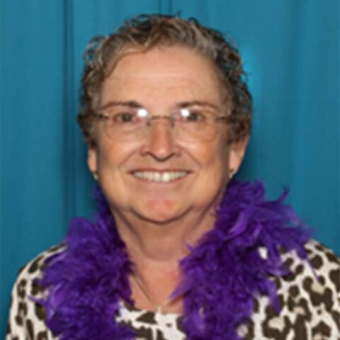 Elderly woman with glasses, smiling, wearing a leopard print blouse and purple feather boa, with a turquoise background.