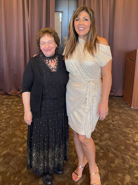 Two women smiling and posing together at an indoor event, one in a black lace dress and the other in a sparkly gold one-shoulder dress.