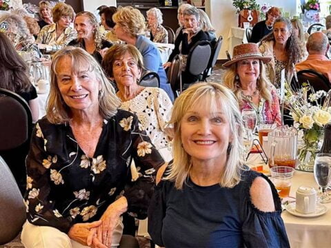 Three smiling women seated at a banquet table with other guests and floral centerpieces in a brightly lit room.
