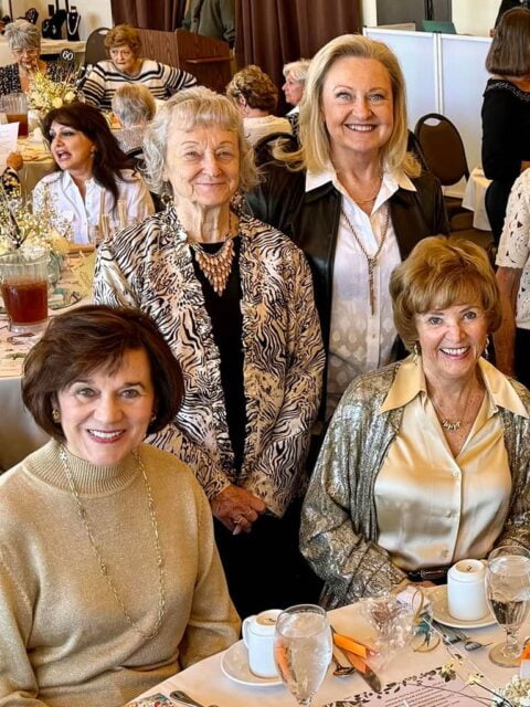 Four senior women smiling at a table during a formal event, dressed in elegant attire with a crowded room in the background.