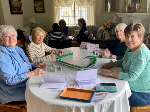 Four elderly women are seated around a table playing a game of Mahjong in a well-lit, cozy room with more people in the background.