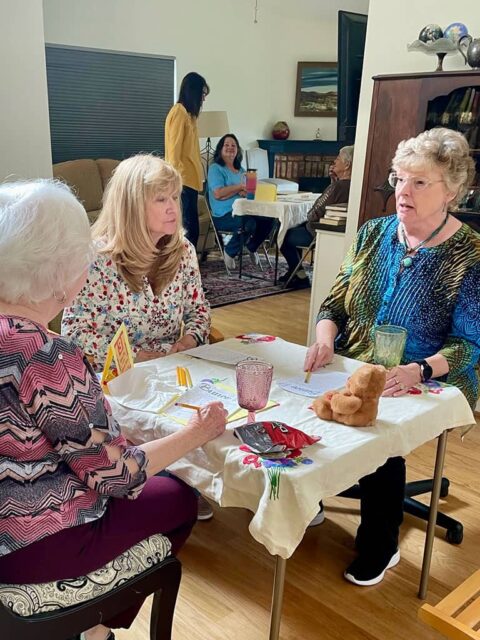 A group of women sit around a table with a teddy bear at home, engaging in conversation. Another person is seated in the background next to a lamp and bookshelf.
