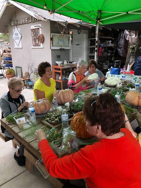 Four women of various ages engaging in crafting activities with plants and pumpkins at a covered outdoor workshop table.