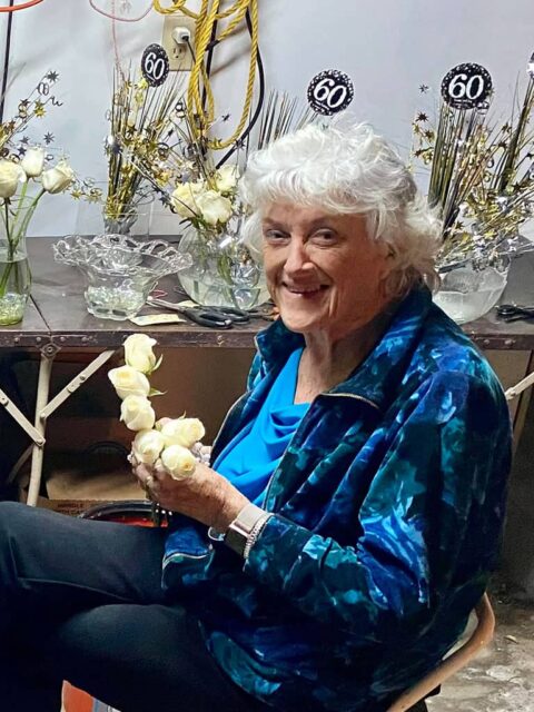 Elderly woman smiling while holding a bouquet of white roses at a celebration with decorations indicating a 60th anniversary.