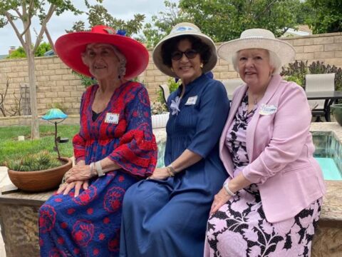 Three women wearing dresses and floral hats sit on a stone bench in a backyard garden. Each wears a name tag. They are smiling and appear to be enjoying a social gathering.