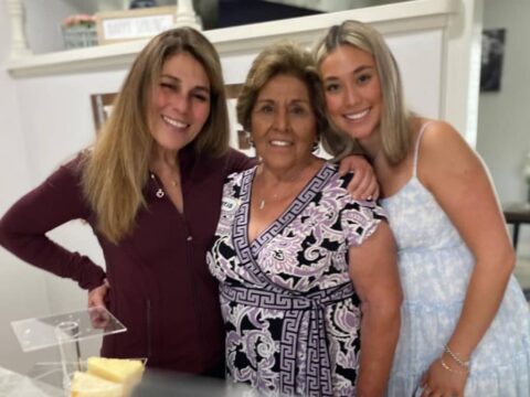 Three women are posing together indoors, smiling at the camera. A piece of cake is visible on the table in front of them.