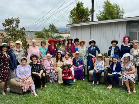 A group of women, many wearing hats, pose for a photo outdoors in a backyard. Some are seated on outdoor furniture while others stand behind them. A building, trees, and power lines are in the background.