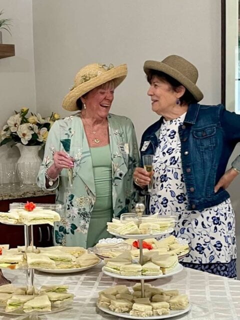 Two women in hats share a conversation while holding drinks at a gathering. A table in front of them is set with plates of sandwiches and floral decorations.