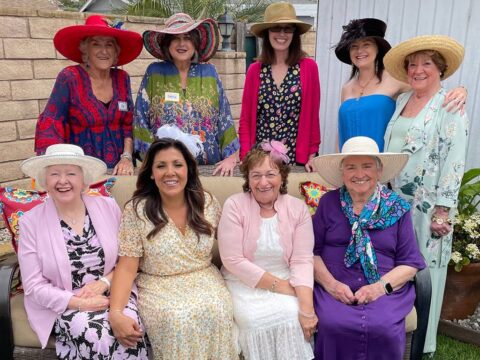 A group of nine women dressed in colorful clothing and various hats pose together outdoors, smiling at the camera. They are arranged in two rows, with some sitting and others standing behind them.