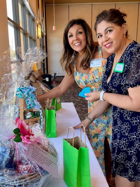 Two women standing at a table, smiling, are distributing or organizing gift bags and items. Several gift bags and wrapped items are on the table in front of them.