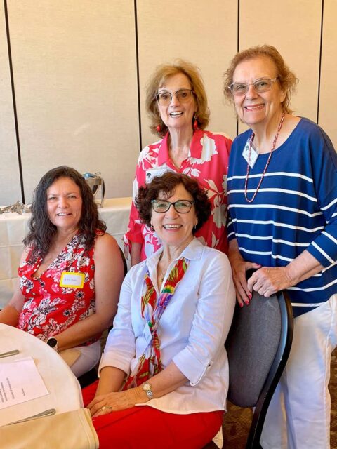 Four women are posing and smiling at a table during an indoor event. Two are seated while the other two are standing behind them. They are dressed in casual attire with vibrant colors.