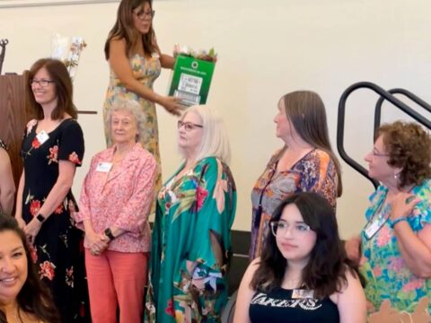 Group of women standing and sitting indoors, some with name tags, one standing woman holds a large green gift bag.