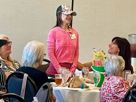 A person in a pink hoodie and baseball cap stands at a table with four seated individuals having a meal. The seated individuals are engaged and smiling.