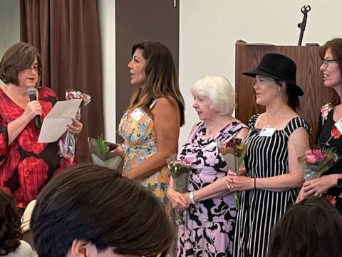 Four women holding flowers stand in a line as another woman, holding a microphone and a piece of paper, appears to be speaking to them.