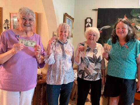 Four women stand indoors holding up dollar bills, smiling. They appear to be in a living room with various decorations on the walls behind them.