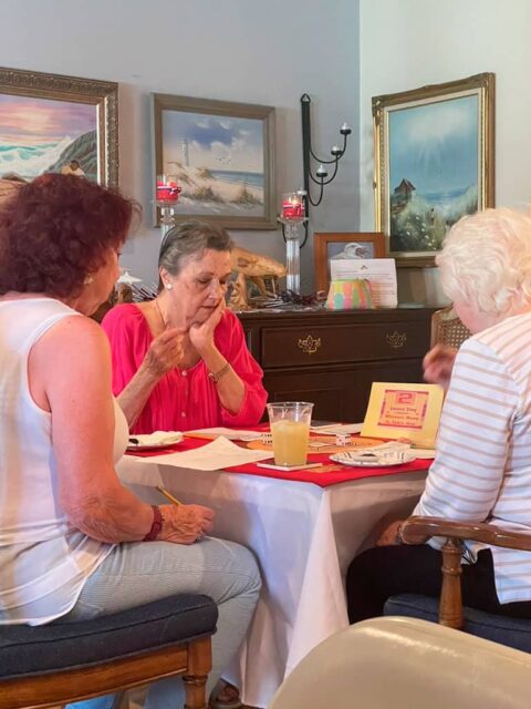 Three women sitting around a table engaged in a card game. The table has cards, a scorepad, and a drink. Paintings and decorative items adorn the background.