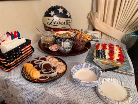 Table with assorted snacks including cookies, fruit, candy, and napkins. Decorative items include a patriotic-themed basket and a "Liberty" pillow. Plates and napkins are set out for serving.