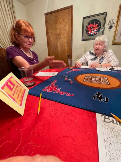 Two elderly women are seated at a table playing Mahjong. The woman on the left is gesturing, and the woman on the right is looking at the game tiles in front of her.