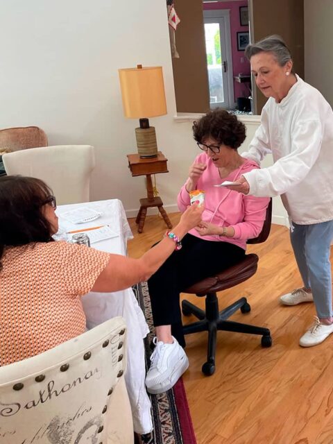 Three women are gathered in a living room. One is seated and pointing to a card, another is showing her something on her phone, and the third is reaching out with a playing card.