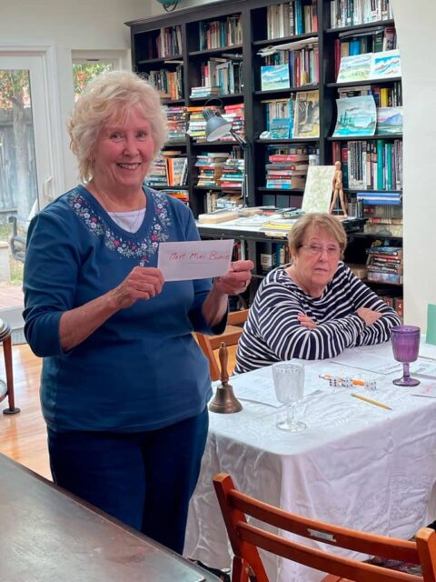 Two elderly women at a table. One is standing and holding a paper with "Not Miss Rosie" written on it, while the other is sitting, looking towards the camera. Shelves filled with books are in the background.