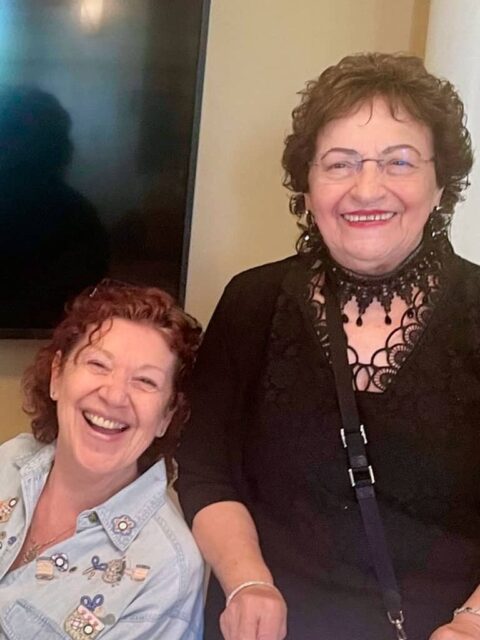 Two women smiling in front of a television. One is seated, wearing a light denim jacket; the other is standing, wearing a black lace top and glasses.
