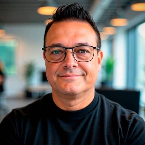 A person with glasses and short dark hair is smiling at the camera in a modern, well-lit office setting.