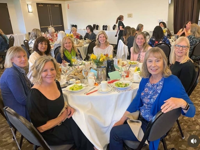 A group of women sit around a round table with food and drinks at a formal indoor event, with more people seated at tables in the background.