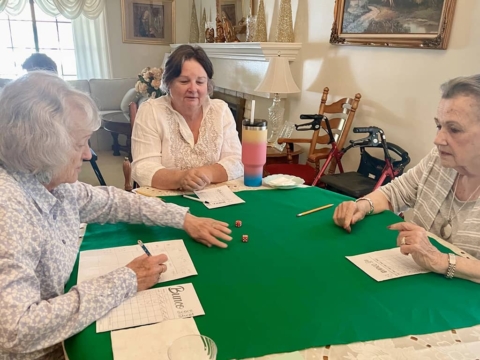 Three elderly women sit around a table with a green cloth, playing a dice game and recording scores on paper sheets.