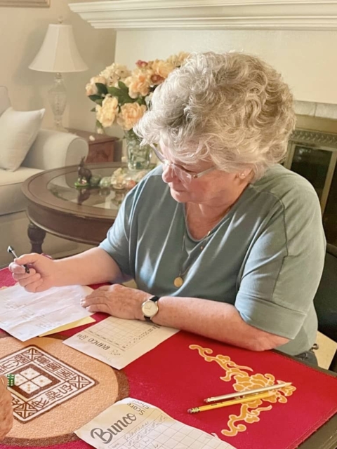 An older woman sits at a table, writing on a score sheet while playing bunco. a glass table with flowers is visible in the background.