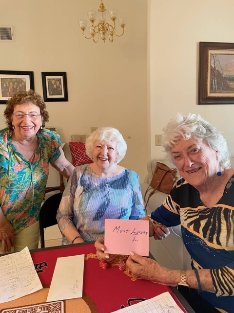 Three older women are smiling at a table. one woman holds a pink envelope labeled “most lessons.” papers and envelopes are spread on the table in front of them.