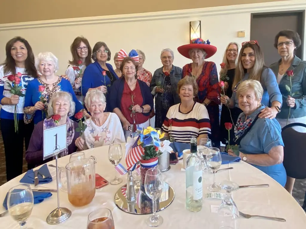 A group of women pose around a table set for a meal, each holding a rose. the table is decorated with american flags and beverages, and some women wear patriotic themed clothing and accessories.