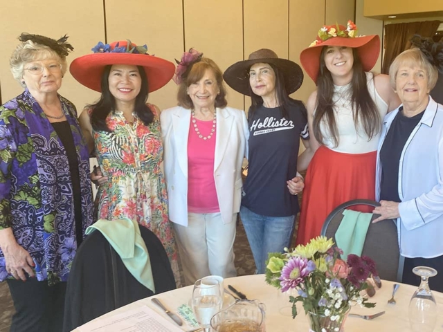Six women stand together indoors, several wearing large decorative hats. a table with flowers, glasses, and napkins is visible in the foreground.