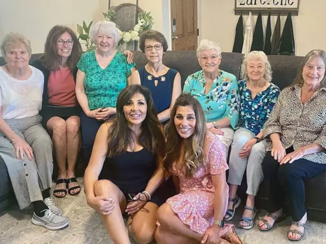 Nine women sit and kneel together indoors, posing and smiling for a group photo on a couch and floor in a well lit room.